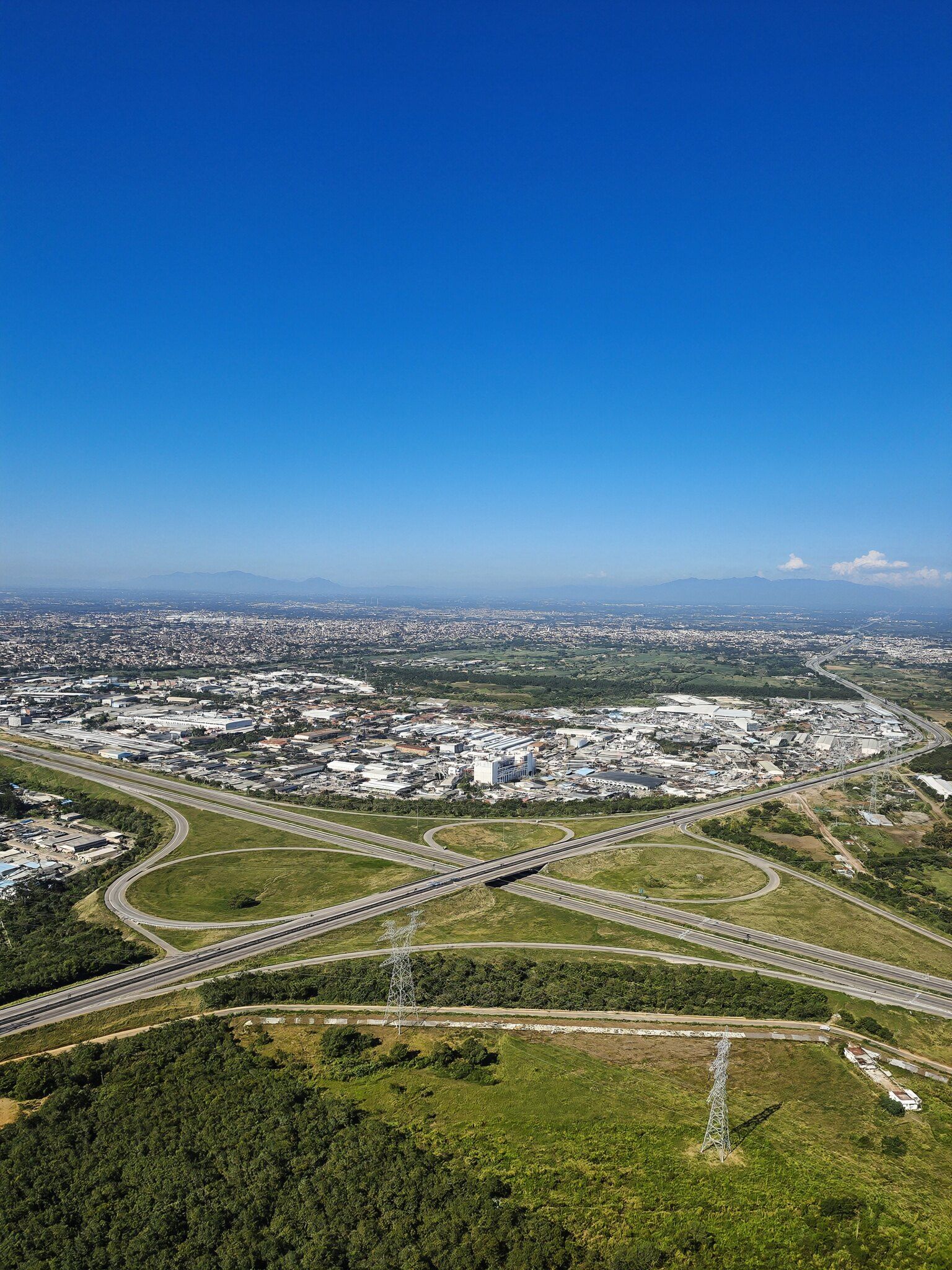 Área industrial ao lado de um trevo rodoviário, com linhas de transmissão cruzando o campo