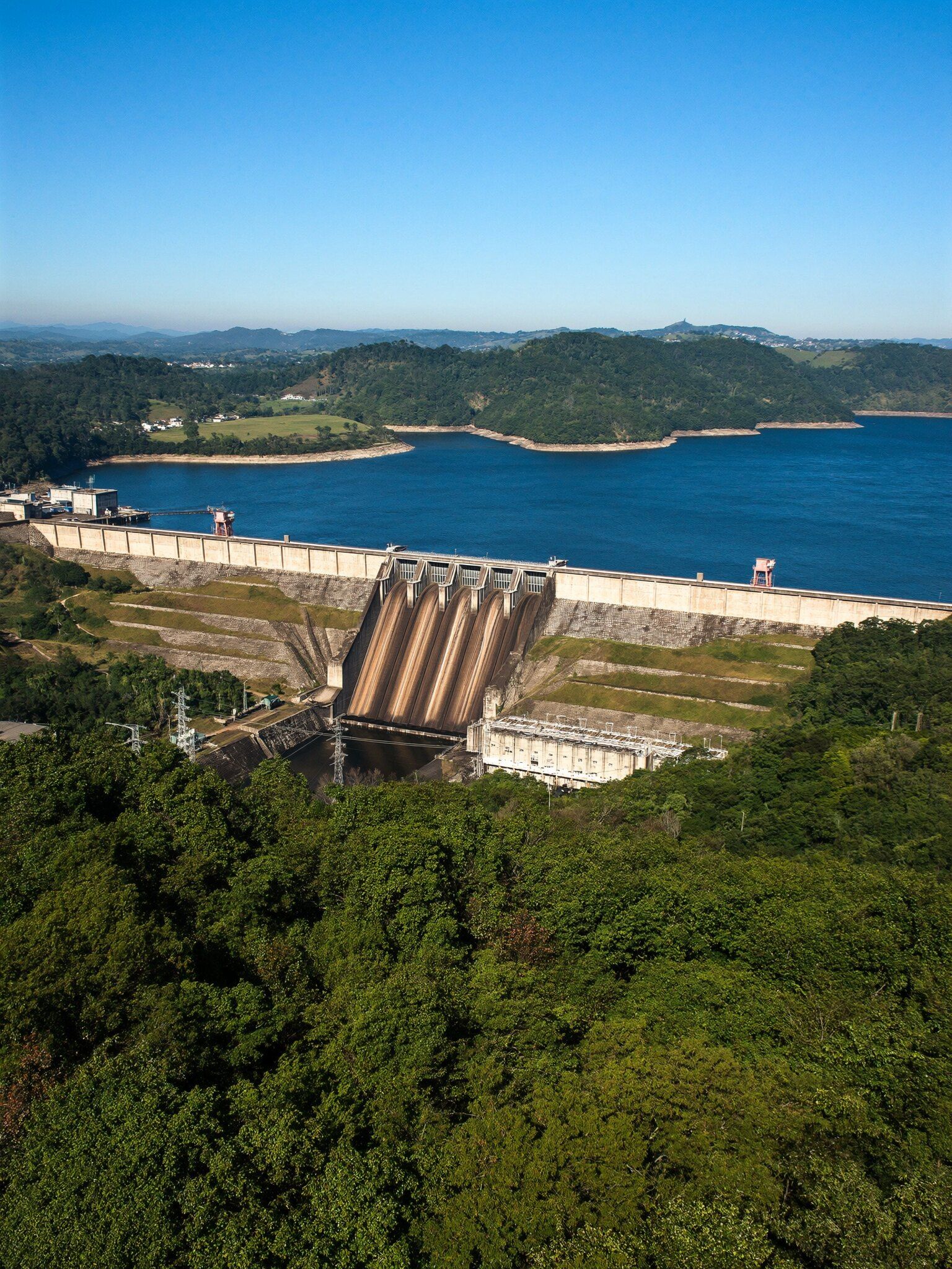 Usina hidrelétrica e represa cercadas de mata, vistas à distância em um dia de sol
