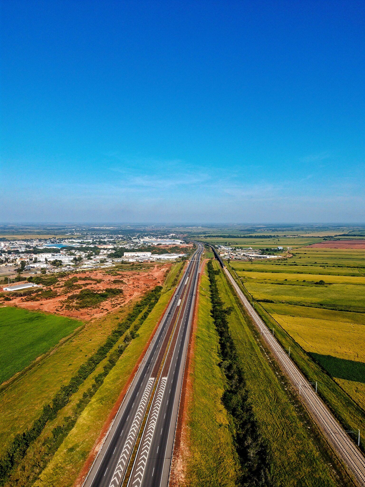 Rodovia e ferrovia cortando lavouras em direção a um distrito industrial, vistas do alto