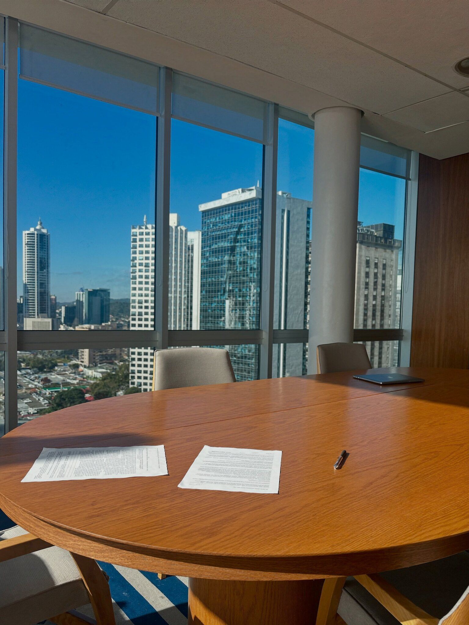 Sala de reunião clara, com documentos sobre a mesa e prédios de escritório ao sol pela janela