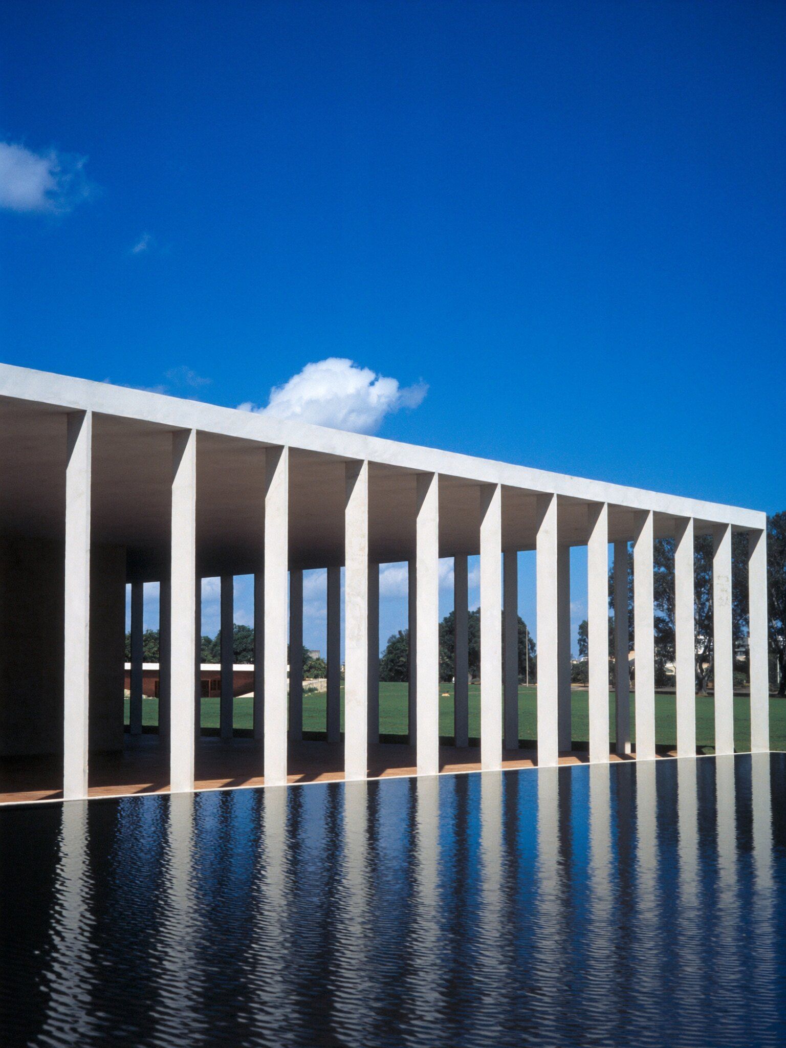 Colunata modernista de pilares brancos refletida em um espelho d’água, sob céu azul